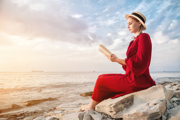 Naklejka na ściany i meble An attractive Caucasian girl in a red bright dress and a straw hat sits on a large stone by the sea at sunset and reads an interesting book on the sea horizon and beautiful clouds.