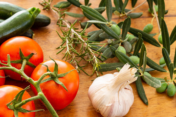 Red tomatoes on branch with zucchini, head of garlic and rosemary twig with green olives on branch with leaves over a used oak wood background. Some of the Mediterranean ratatouille recipe ingredients