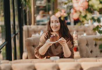 Charming brunette woman with long curly hair sitting at window in cafe with dessert in hands, selective focus