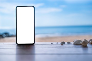stock photo phone in the beach, black smartphone show white screen placed on wooden table with shells. Sky summer,beach and sea in background with copy space , blank for text ads and graphic design.