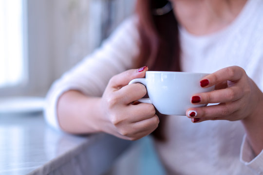 Closeup beauty portrait model hands with red fashion nails painting in warm sweater holding a white cup of coffee, tea, milk or another beverage in morning next to window at home, mockup for designs.