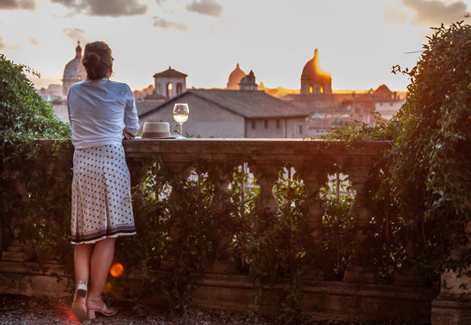 Young Woman Tourist Fashion White Dress With Glass Of White Wine In Front Of Panoramic View Of Rome Cityscape From Campidoglio Terrace At Sunset. Landmarks, Domes Of Rome, Italy.