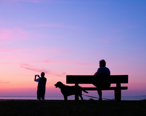 Fototapeta na wymiar man takes pictures of colorful sunset while woman sits with dog on bench