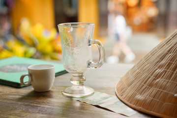 Empty tall transparent glass of macchiato or capuccino coffee with small white syrup mug and Vietnam money banknote and Vietnamese straw hat with traditional hat in coffee. Shop as background