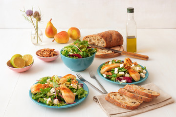 White wooden table with walnut salad, caramelised pears and feta cheese. Turquise dishes with salad, slices of twisted swiss bread with olives, limets, bottle of olive oil and whole pears.