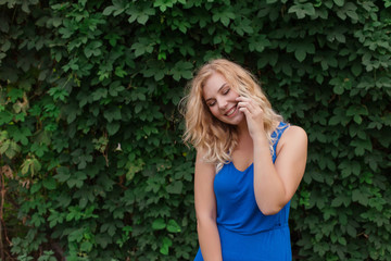 Beautiful young girl in a blue dress talking on the phone. Against the background of wild grapes, summer day. Copy space.