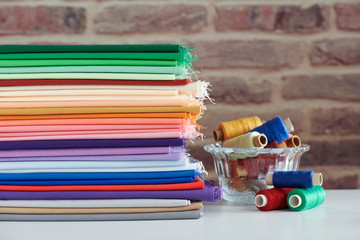 Fototapeta premium Stack of colorful monochromatic fabrics and spools of thread on brick wall background