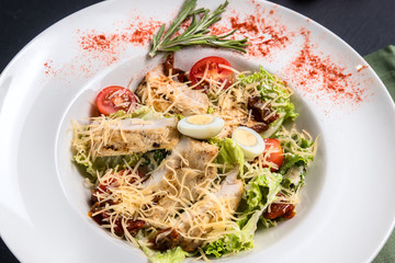 Close-up view on classic Caesar salad with grilled chicken breast and half of quail eggs in white ceramic plate. Green tablecloth, spoon and fork on dark gray texture background