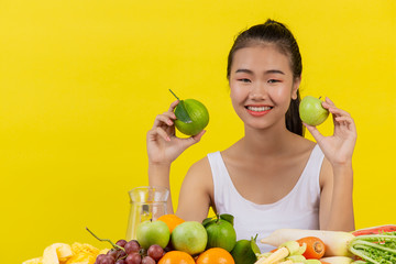 An Asian woman wearing a white tank top. The left hand holds an apple, the right hand holds an orange and the table is full of many fruits.