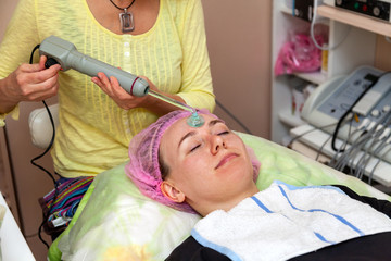 Obraz na płótnie Canvas A young girl is lying on couch with a pink hat on her hair while performing an darsonvalization on face with a special device while cleaning and removing body fat under the skin on nose. Cosmetology