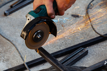 Close-up view strong man master without gloves on arms, performs metal cutting with an angle grinder in the garage workshop, blue and orange sparks fly to the sides