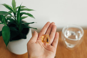 Dietary supplements. Hand holding omega 3 two capsules above wooden table with glass of water....