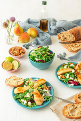 White wooden table with walnut salad, caramelised pears and feta cheese. Turquise dishes with salad, slices of twisted swiss bread with olives, limets, bottle of olive oil and whole pears.