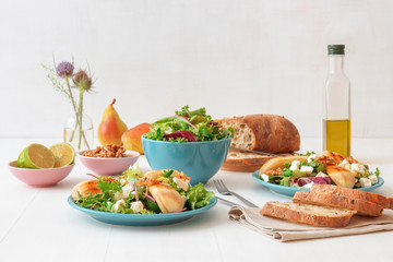 White wooden table with walnut salad, caramelised pears and feta cheese. Turquise dishes with salad, slices of twisted swiss bread with olives, limets, bottle of olive oil and whole pears.