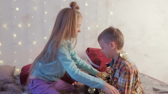 Boy and girl children friends playing with garland sitting on bed at home in Christmas interior. Cousin, siblings, child friendship. Celebrating new year xmax holiday, fun pastime in childhood.