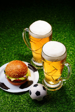 Two glasses of beer, hamburger and soccer ball on green grass.