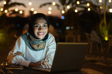 Smiling business asian muslim woman browsing on laptop at outdoor cafe in evening.