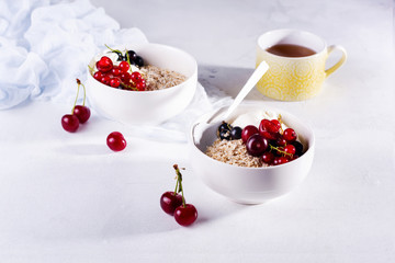 Raw oatmeal with yogurt and berries of cherry, currant and blueberry for making healthy breakfast in white ceramic bowl on a light background. Selective focus.