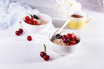 Raw oatmeal with yogurt and berries of cherry, currant and blueberry for making healthy breakfast in white ceramic bowl on a light background. Selective focus.