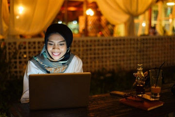 Smiling business asian muslim woman browsing on laptop at outdoor cafe in evening.