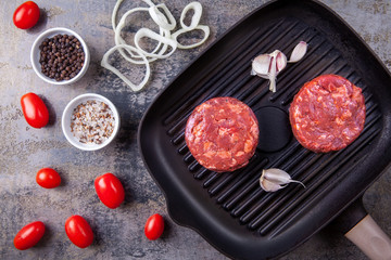 Ready to cook two raw beef burger meats with garlic on a grill pan, cherry tomatos, onion rings, colored salt and pepper on a gray marble background. Recipe. Top view.
