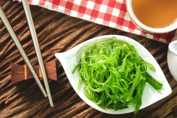 Hiyashi Wakame Chuka or seaweed salad in white bowl leaf with stainless steel chopsticks and teacup on wooden table, Japanese food