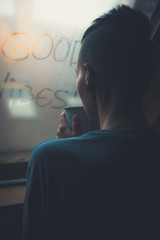 Young girl with short, freshly raised hair drinks coffee looking out the window. The window is fogged and "good vibes" is written on it.