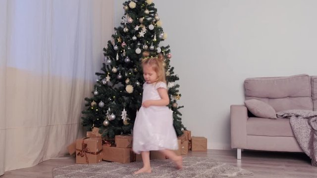 Barefooted little cute girl in beautiful white dress dancing near Christmas tree at home in living room. Traditional New Year interior. Winter xmas holiday.