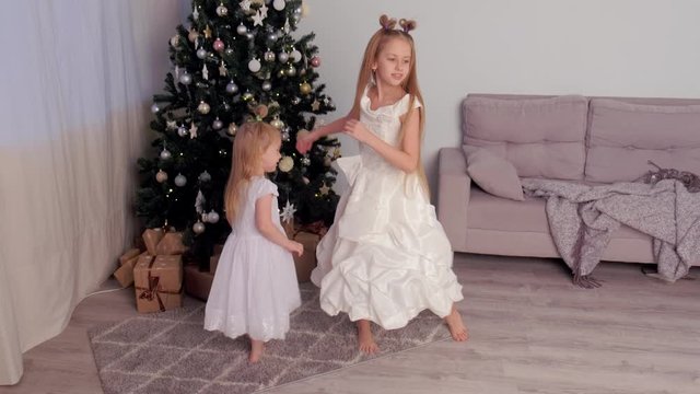 Two sisters children girls in beautiful white dresses whirling and dancing near Christmas tree at home in traditional xmas interior in living room. Family time at winter New Year holidays.