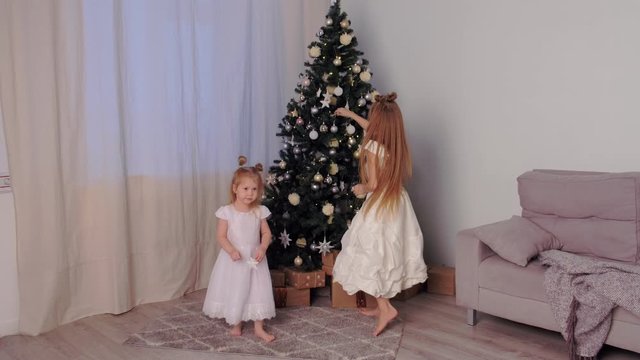 Two sisters in beautiful white dresses whirling and dancing near Christmas tree at home in traditional xmas interior in living room. Family time at winter New Year holidays.