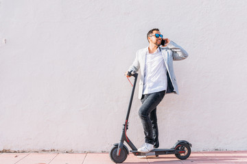 Latin adult man with sunglasses, well dressed and electric scooter talking on his mobile phone sitting on the street with a white wall background © pedro