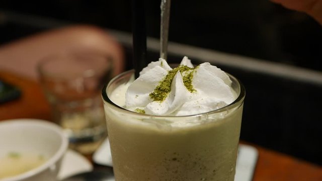 Delicious matcha tea frappe with whipped cream in glass cup. Girl eats cream with a spoon in glass with matcha iced tea. CloseUp