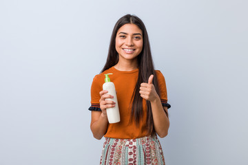 Young arab woman holding a cream bottle smiling and raising thumb up