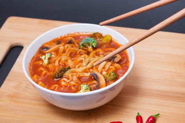 Bol de noodles veganos picantes con verduras sobre una tabla de madera con guindillas y palillos de madera sobre fondo negro.