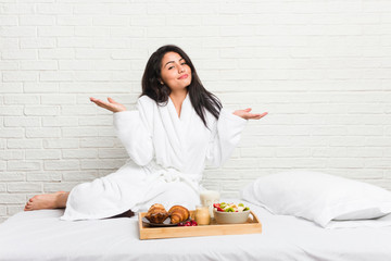 Young curvy woman taking a breakfast on the bed doubting and shrugging shoulders in questioning gesture.