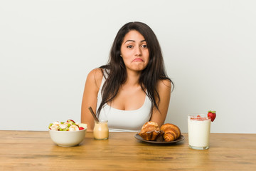 Young curvy woman taking a breakfast shrugs shoulders and open eyes confused.