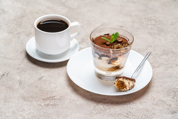 Portion of Classic tiramisu dessert in a glass and cup of coffee on concrete background