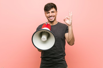 Young hispanic man holding a megaphone cheerful and confident showing ok gesture.