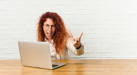 Young redhead curly woman working with her laptop showing a horns gesture as a revolution concept.