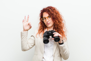 Young caucasian business redhead woman holding a binoculars cheerful and confident showing ok...