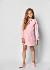 Beautiful little girl with long blond hair and blue eyes, standing on a white background, wearing a pink casual dress and gym shoes, teenager style