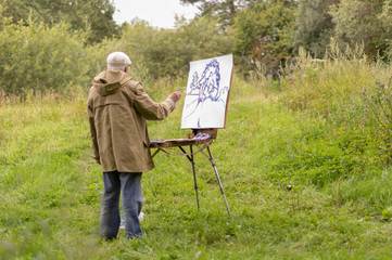 An elderly man is interested in painting with oil paints. Drawing a natural landscape. On white cardboard