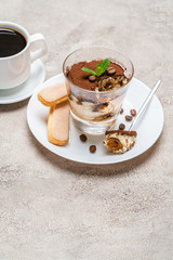 Portion of Classic tiramisu dessert in a glass, savoiardi cookies and cup of coffee on concrete background