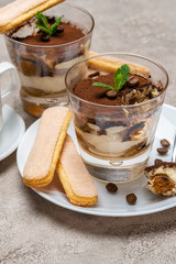 Portion of Classic tiramisu dessert in a glass, savoiardi cookies and cup of coffee on concrete background