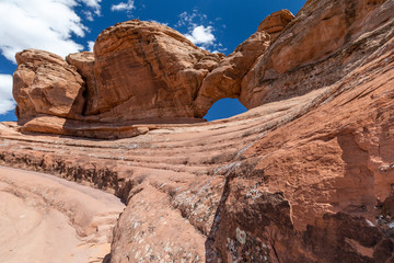 Wide angle view of an imposing red sandstone rock formation with a natural arch on top, under a blue sky with puffy clouds