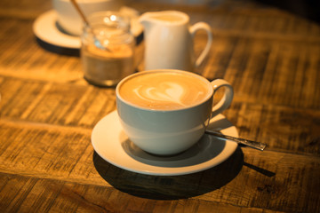 A cup of coffee latte with heart shaped decoration from cream on the wooden table in a cafe. Glass pot with brown sugar and tiny white milk jar in the background.