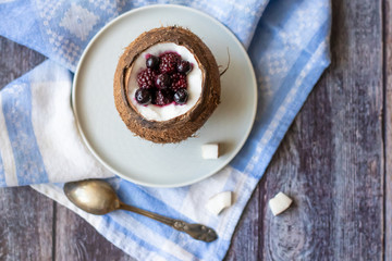 Berry dessert with yogurt in a cup of coconut on a wooden background. Blackberries, raspberries, blueberries, black currants. Blue napkin