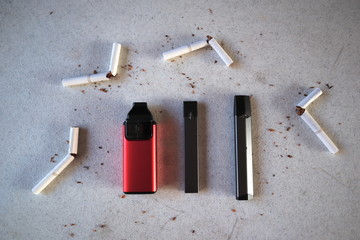 Different vape devices electronic cigarettes as smoking alternatives with broken cigarettes and scattered tobacco on white textured background