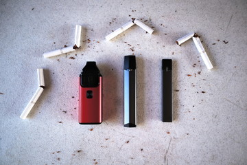 Different vape devices electronic cigarettes as smoking alternatives with broken cigarettes and scattered tobacco on white textured background