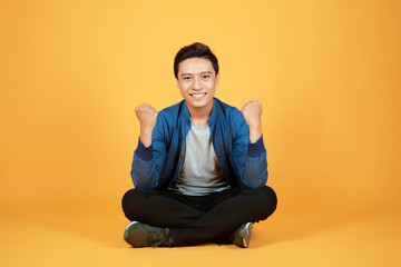 Handsome cheerful young Asian guy wearing casual jeans denim, shoes, sitting on floor with crossed legs, raising hands up. Isolated on orange background